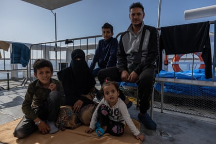 Two children and a woman in a burqa sit on the deck of a ship, while a man and another child sit on a bench next to them.