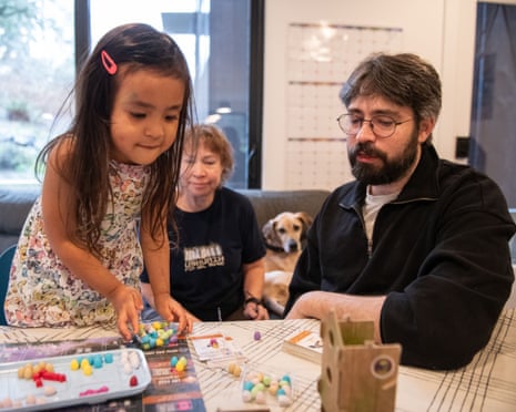 Young girl and man sit at a table with a board game, as woman and dog look on from couch behind them.