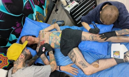 Top-down shot of two tattoo artists working on pieces on a man’s arm and leg