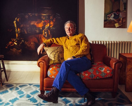 Guy Kennaway at his home, wearing bright blue jeans and a mustard-coloured top, leaning back on a sofa with a huge painting behind him