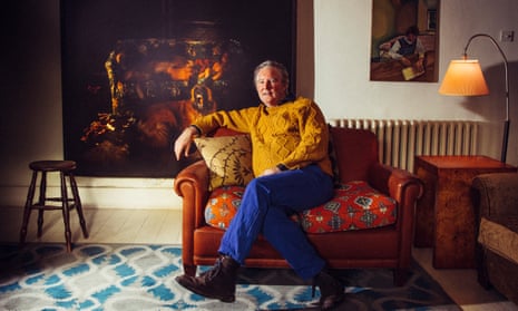 Guy Kennaway at his home, wearing bright blue jeans and a mustard-coloured top, leaning back on a sofa with a huge painting behind him