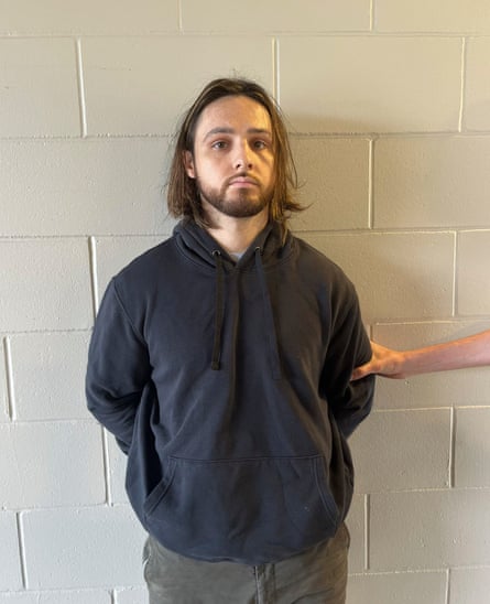 A man, unsmiling, in a black hoodie, goatee, and shoulder-length brown hair, looks at the camera as an arm holds him against a white cinderblock wall from the .eft