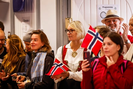 Men and women wave Norwegian flags. One blond woman wears traditional Norwegian costume with a white blouse and red waistcoat.