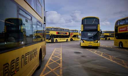 Bright yellow double decker buses with Bee Network logos and branding are seen at the Bolton Interchange; they are stationed and manoeuvring around parking bays marked out with yellow paint, and their colour contrasts with the grey tarmac and cloudy blue sky