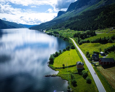 A narrow road runs along a lake with mountains sloping down to the water and clouds reflected in it
