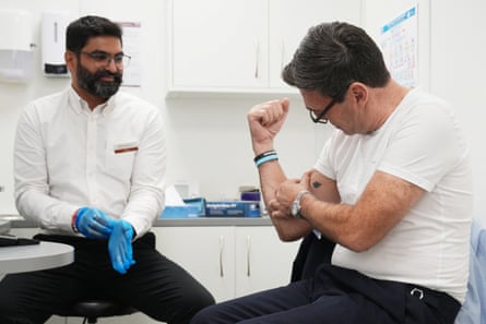 Andy Burnham in a T-shirt flexing his arm for the benefit of a doctor