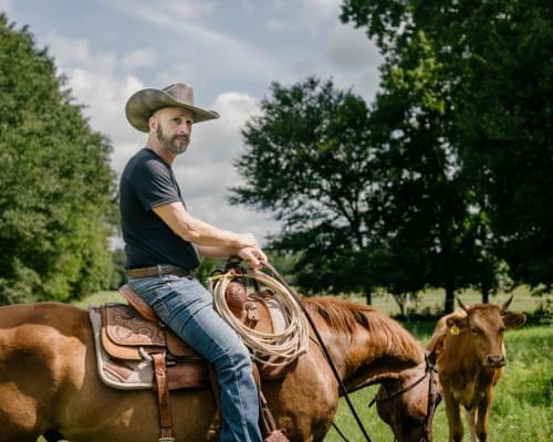 Michael Samadi riding a horse in a large area of green grass, near a tree where a herd of horses are standing in the shade