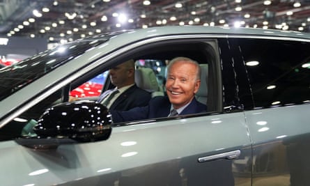 A smiling older man wearing a suit sits in the driver’s side of a car, with a larger man in a suit looking straight out the windshield, unsmiling (likely Secret Service). They are inside a large hangar with rows of lights on the high ceiling.