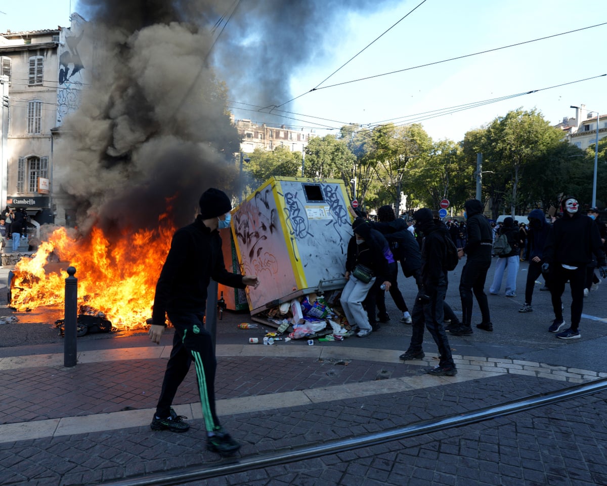In Marseille, a person walks past a burning dumpster as masked protesters tip over a bin.