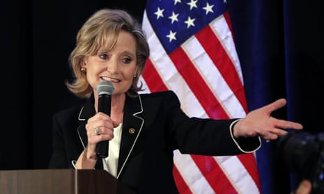 Middle-aged blond white woman in black blazer smiles holding microphone with one hand and gesturing beyond lectern with the other, in front of American flag.