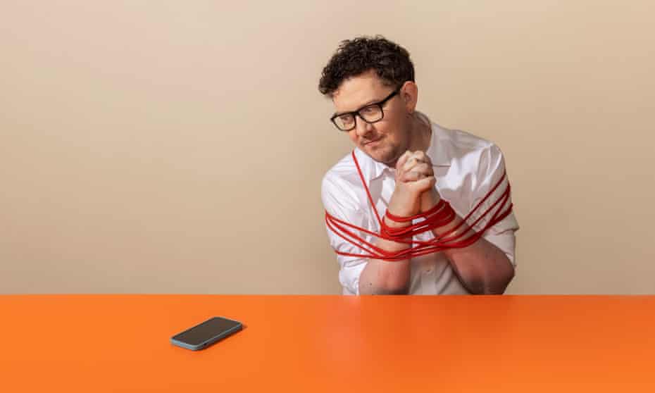 James Ball looking longingly at a smartphone on an orange desk, with his hands tied together in front of him