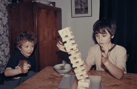 Two boys seated a a table look on as a pile of wooden blocks collapses