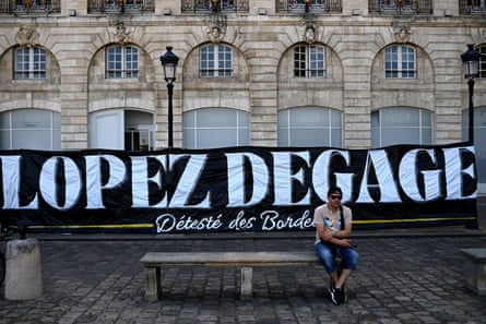 A man sits on a bench in front of a banner reading ‘López get out’ in Bordeaux in May.