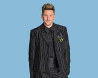 Henry Holland wearing a crinkly black satin suit with a black cummerbund and bow tie, smiling at the camera, against cornflower-blue background