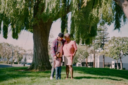 A woman stands with her arm around a man under a large willow tree. A small girl stands between them.