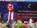 poster of man in a suit is painted with red X over his face by a memorial site with flowers and flags