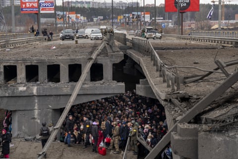 People crowd under a destroyed bridge with vehicles above