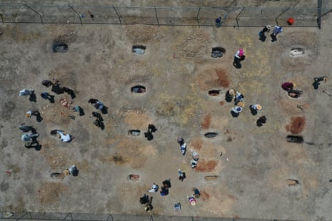 Drone view of about 30 people standing around 16 grave-sized holes in the ground