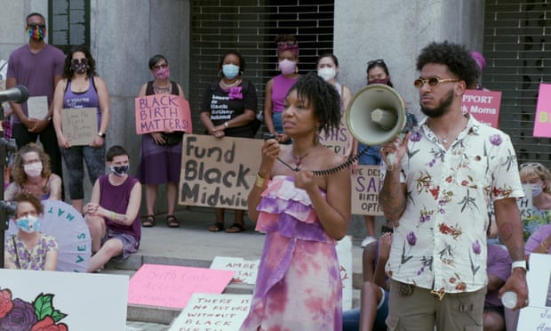 A man holds a megaphone while a woman standing next to him speaks into the handset attached to the megaphone. Behind them, a group of people wearing protective masks hold signs