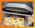 A pizza topped with cheese, peppers, and tomatoes sits before an open Ooni pizza oven