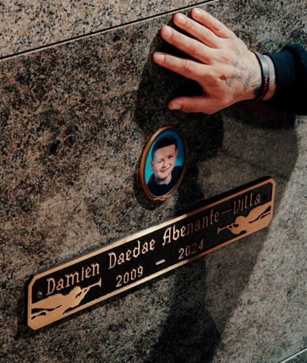 Alex Villa’s hand is shown against the headstone of his son’s grave, which has a small circular photo of Damien on it and a name plaque