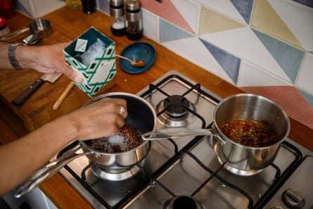A closeup of a man’s hands over a hob with two saucepans with oil and spices in them