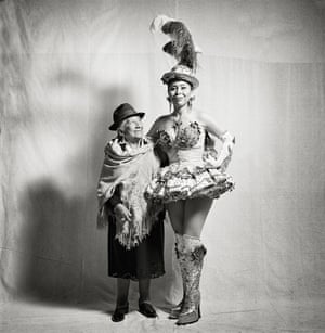 An old Bolivian woman in a shawl smiling up at her companion, a tall woman in chorus-line outfit and feathered hat