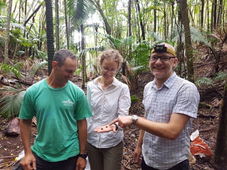 Three people in a native forest, smiling and looking at two tiny snails in the hand of the woman standing in the middle.
