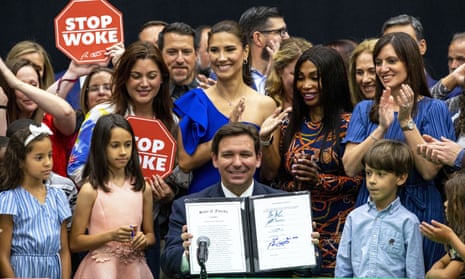 desantis smiles as he holds up bill, surrounded by cheering people. some have signs that say 'stop woke'
