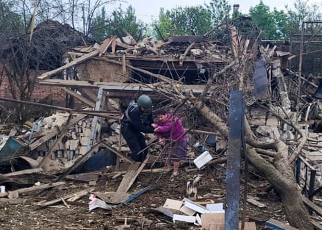 Person wearing helmet helping woman walk from completely destroyed home.