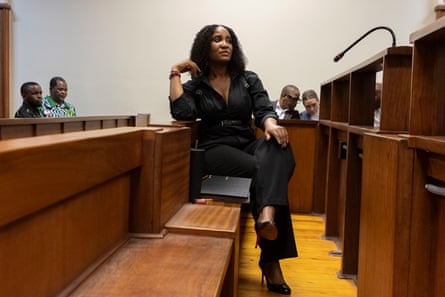 دودوزيل زوما-سامبودلا تظهر في المحكمة: تجلس على مقعد خشبي وترتدي بذلة سوداء ضيقة مع كعب عالٍ رفيع. هي في أوائل الأربعينات من عمرها وشعرها المجعد بطول الكتفين منسدل.