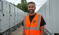 Amit Gudka stands smiling for a photograph in a hi-vis vest outdoors, with two banks of electrical equipment behind him