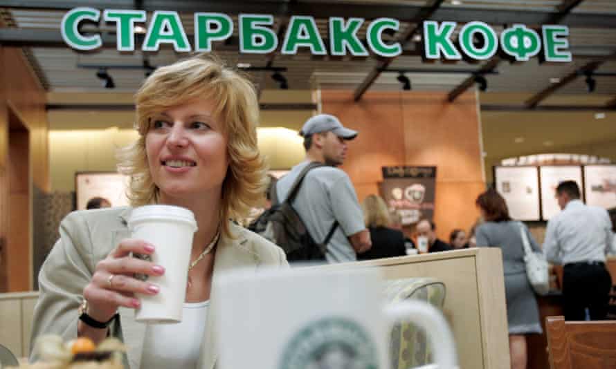 A young woman drinking coffee at a table with the familiar green Starbucks corporate lettering behind her, except the lettering is in Cyrillic rather than the Roman alphabet