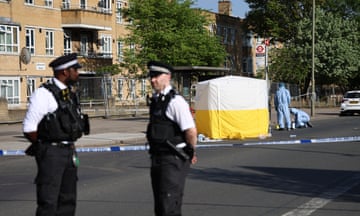 The area of the attack at Burnt Oak Broadway, Edgware: two Met officers stand in front of police tape sealing off the street while forensics officers in pale blue overalls search the scene; a yellow and white tent has been erected at the bus stop, which is in front of a block of flats.