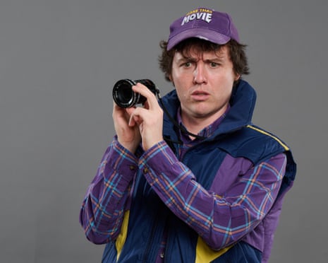 A young man holding a camera wearing a purple plaid shirt and cap and blue gilet. Looking confused into the middle distance