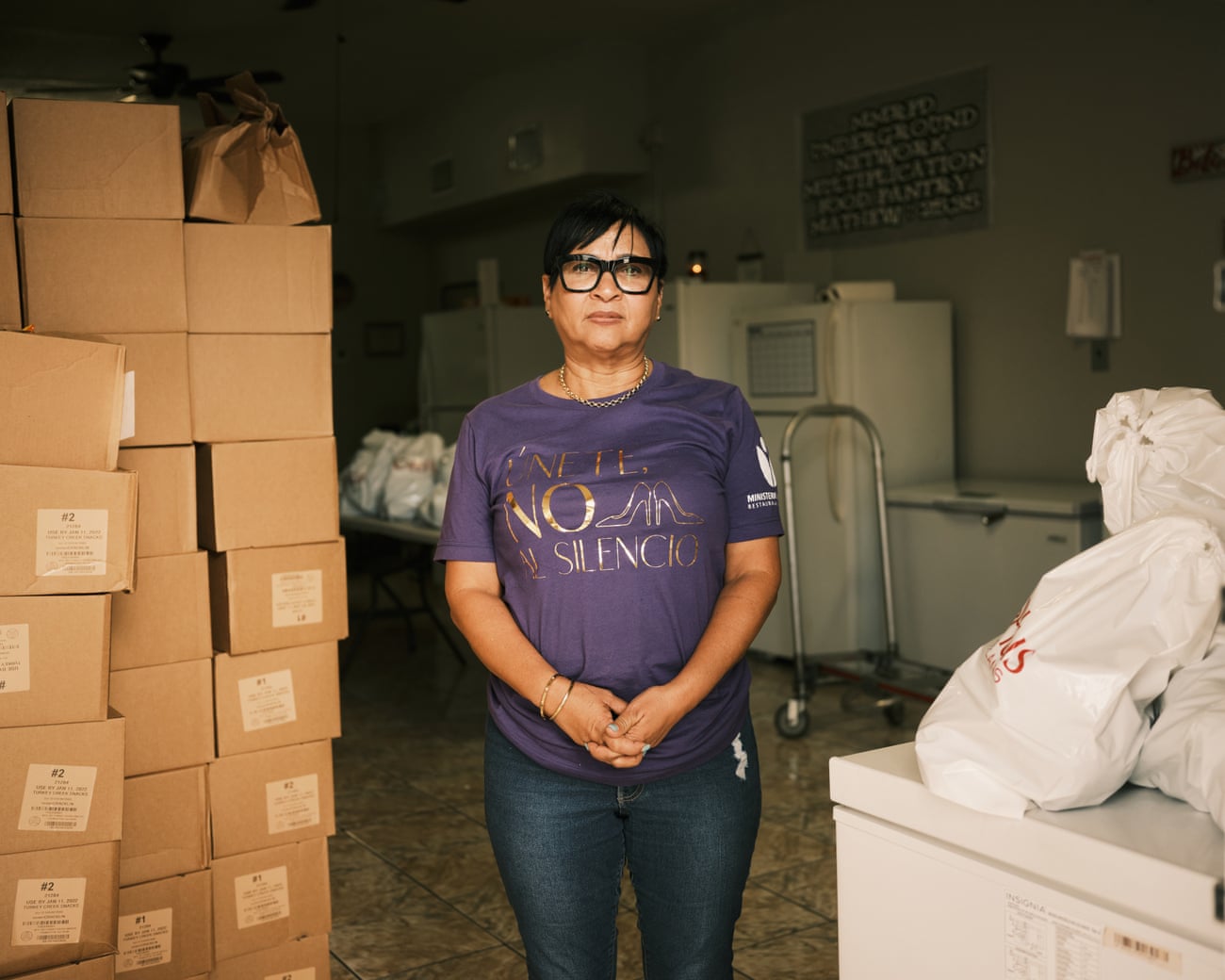 A woman in glasses and a purple shirt stands in the portrait next to several stacks of boxes taller than her height.