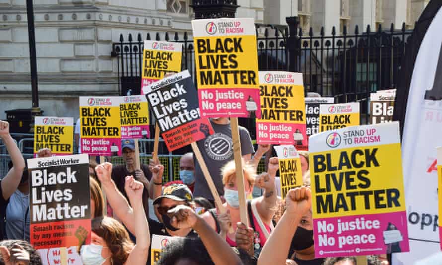 A group of people carrying a lot of yellow, pink and black Black Lives Matter placards close together, with the iron railings of Whitehall or Downing Street behind