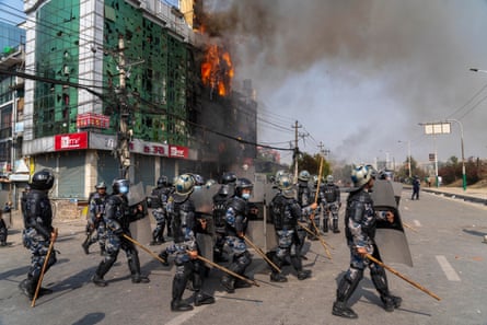 Police with riot shields guard a tall building which has flames and smoke pouring from an upper floor