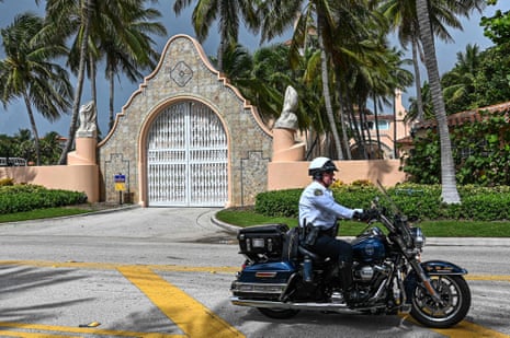 A local law enforcement officer drives a motorcycle past the pink gates of Mar-a-Lago, which is surrounded by palm trees and greenery.