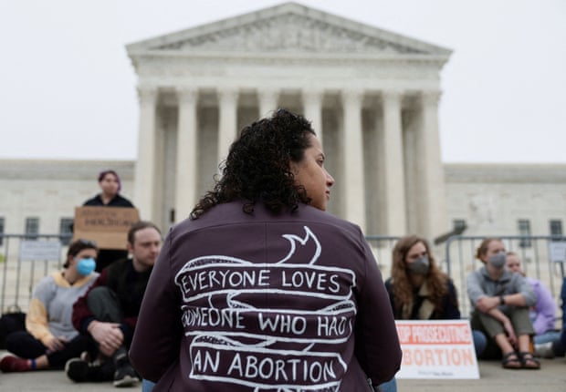 A woman sits facing the supreme court building, her back to the camera. She's wearing a shirt with a message on the back that reads 'Everyone loves someone who had an abortion'.
