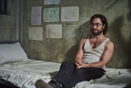 A bearded man sitting on a bed, wearing a white singlet and black trousers.
