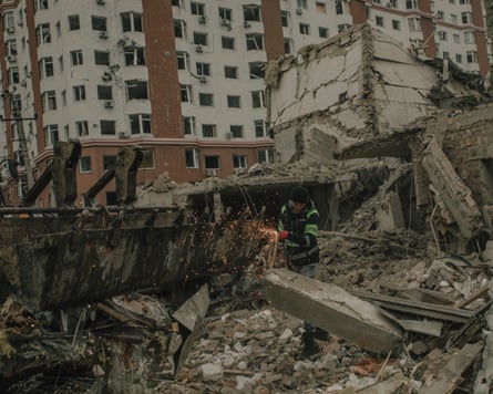 A welder repairs a digger surrounded by rubble, with a block of flats in the background
