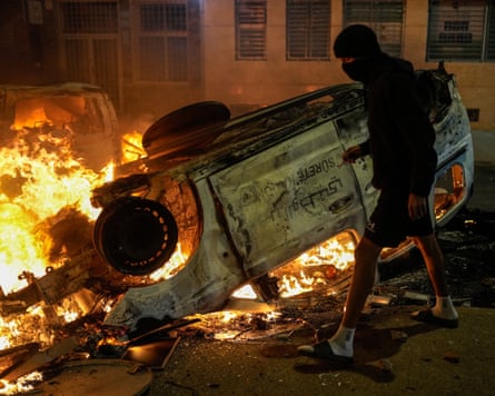 A person wearing a balaclava walks in front of an overturned burning police car