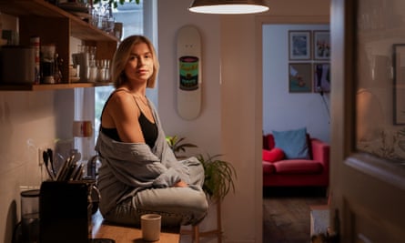 Daisy Hyde, in jeans and a shirt pulled down over her vest, sits cross-legged on a kitchen counter with a sofa in a room behind her