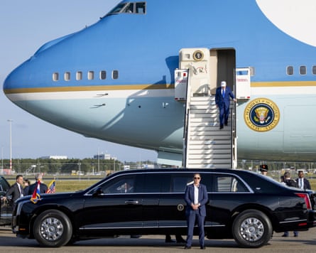 Trump steps down from the large blue-painted plane to a waiting black limousine. He is wearing a white baseball cap with the letters USA.