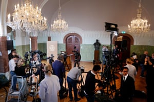 Journalists wait around with cameras in a room with chandeliers and an empty lecturn