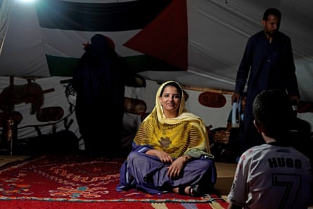 Asria Mohamed, smiling, sits cross-legged on rug on floor in front of Sahrawi flag with people around her