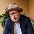A man with stubble wearing glasses, a brown hat, a white T-shirt and a navy jacket