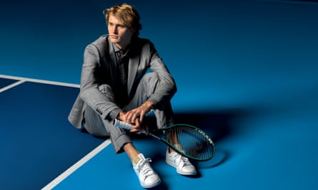 Alex Zverev in a shiny grey smart-casual suit and shirt, and white trainers, holding a racquet, sitting on the edge of a tennis court.