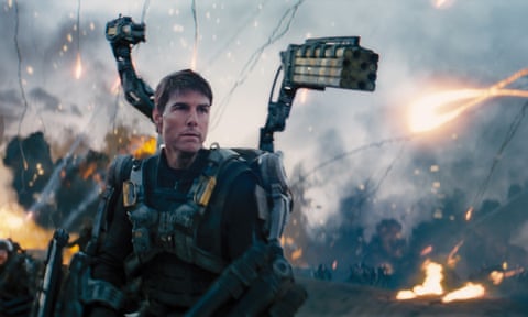 Tom Cruise wearing a mechanical battle suit, standing on an active battlefield with patches of fire around him.